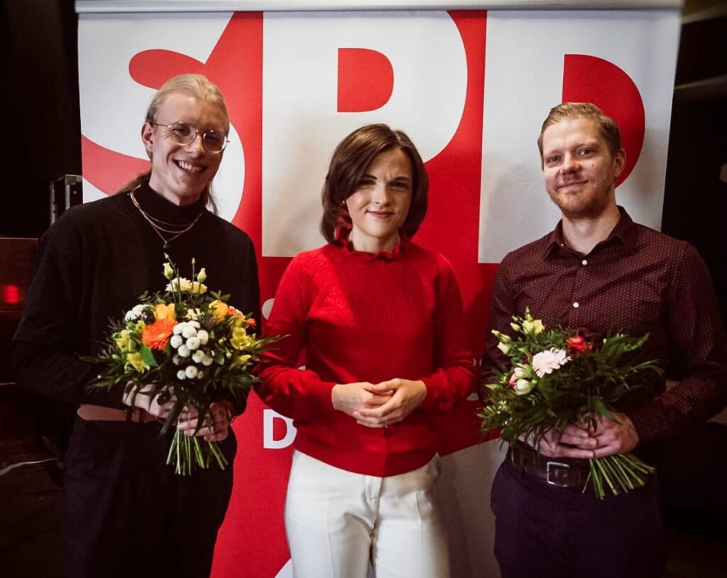 Drei Personen nebeneinander.1. Person weiblich (Mana Klötzer), 2. Person weiblich (Elisabeth Kaiser Bundestagsabgeordnete) und 3. Person männlich (Christian Urban) Mana Klötzer und Christian Urban halten einen Blumenstrauß in den Händen, den sie von Elisabeth Kaiser erhalten haben.