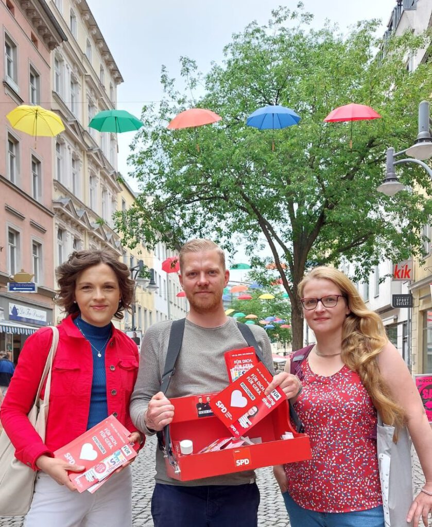 Hintergrund: Kulisse Sorge Gera mit hängenden bunten Regenschirmen. Front drei Personen: Elisabeth Kaiser Bundestagsabgeordnete, Christian Urban, Janine Puschendorf